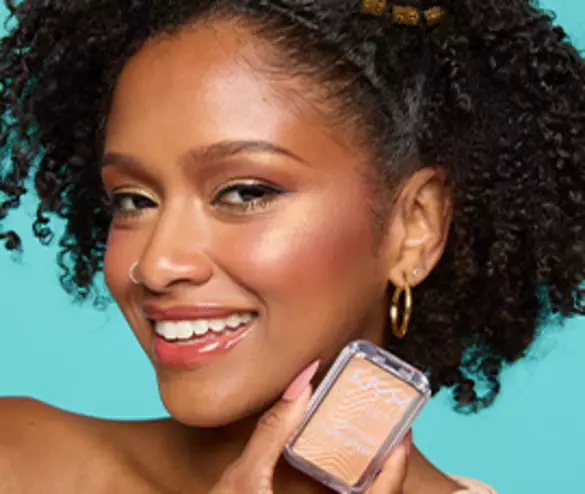 A smiling woman with curly hair and gold accessories holds a NYX makeup product against a bright teal background.