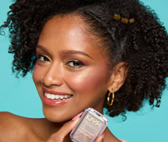 A smiling woman with curly hair holds a makeup product against a bright turquoise background.