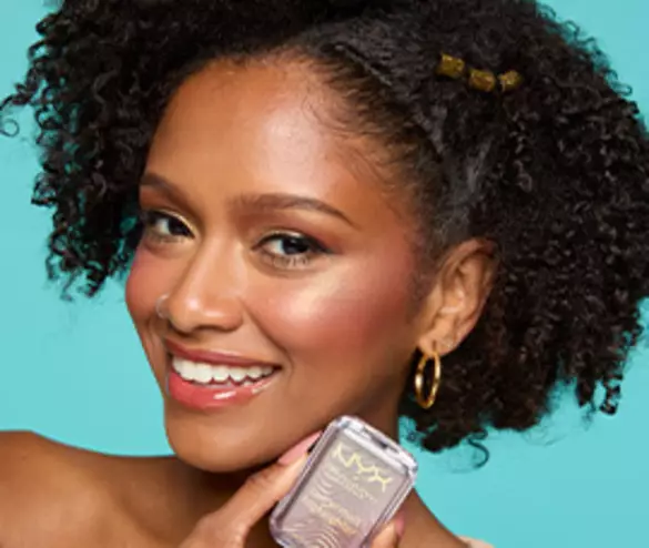 A smiling woman with curly hair holds a makeup product against a bright turquoise background.