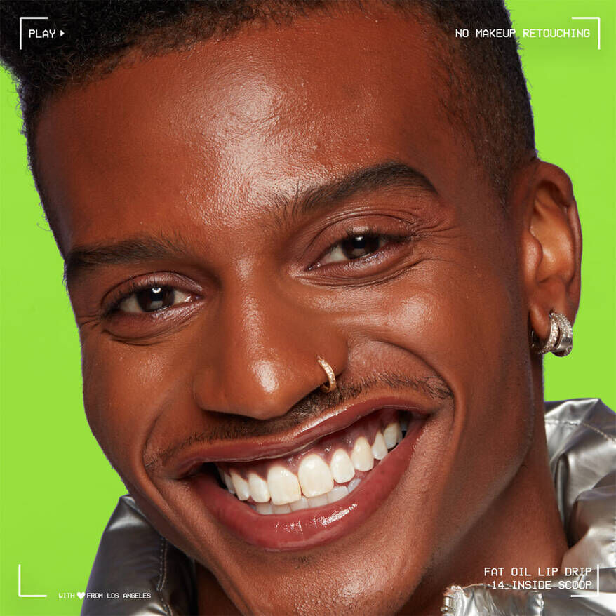 A close-up of a smiling young man with a nose ring, showcasing natural skin and glossy lips, wearing a shiny silver jacket.